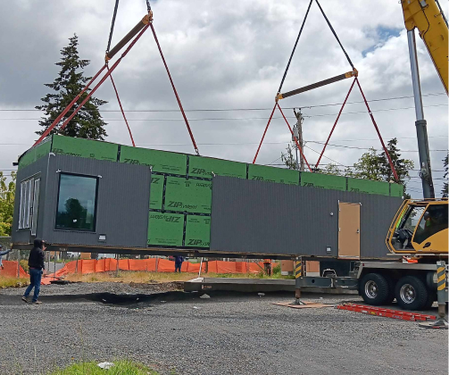 A large modular building section is being lifted by a crane at a construction site, with construction workers observing.