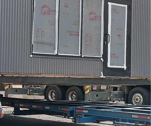 A partially constructed building is on a flatbed trailer. The structure has a door and windows covered with protective wrapping.