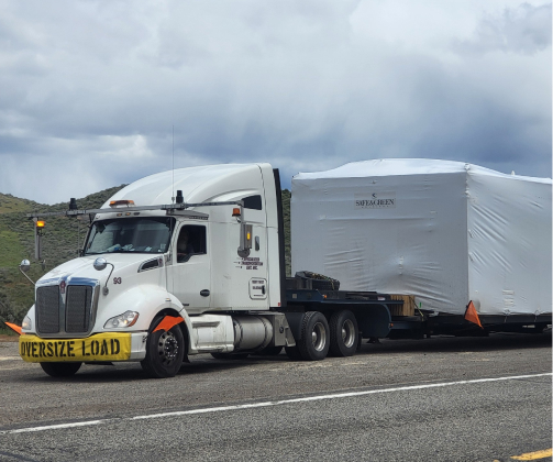 A truck with an "Oversize Load" sign is transporting a large, wrapped object on a flatbed trailer on a road. The sky is cloudy.