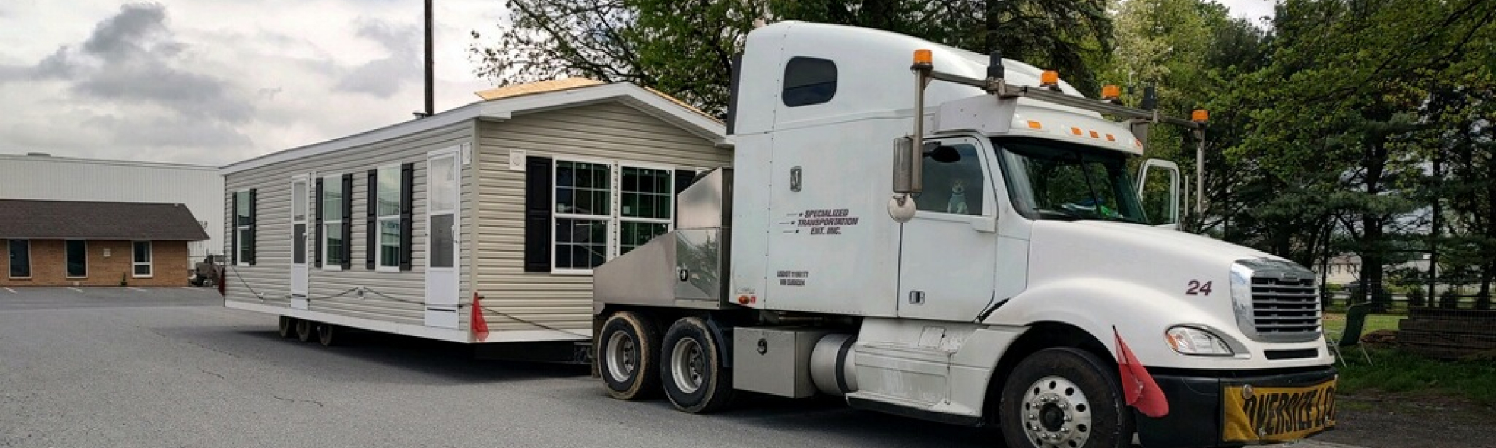 A white semi-truck transports a modular home on a trailer through a parking lot with trees and a building in the background.