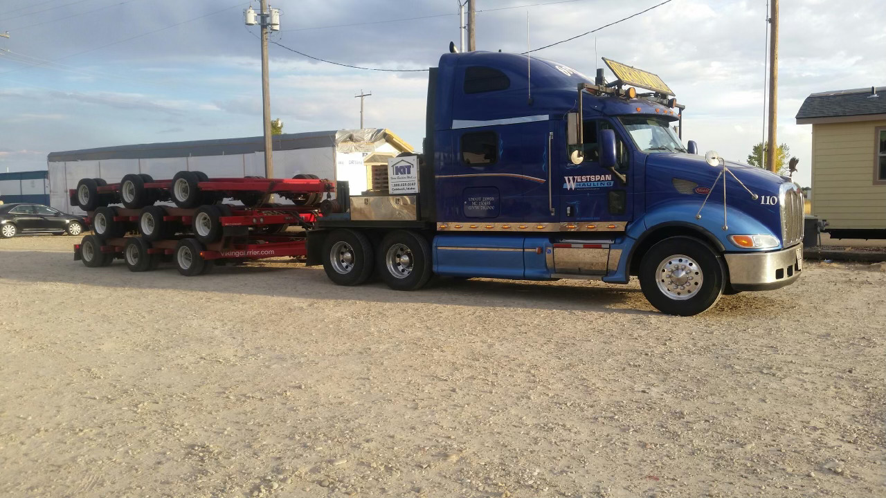A blue semi-truck with a long flatbed trailer transporting several red axles and machinery on a gravel lot.