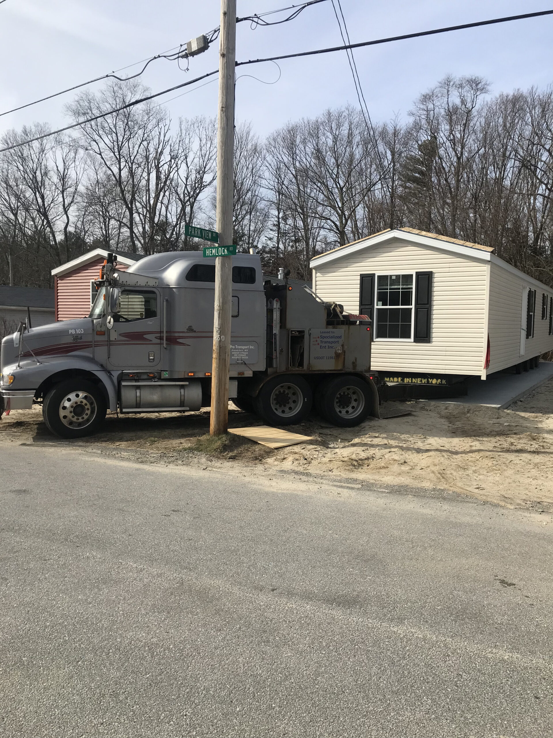 A large truck is transporting a mobile home down a residential street lined with leafless trees.