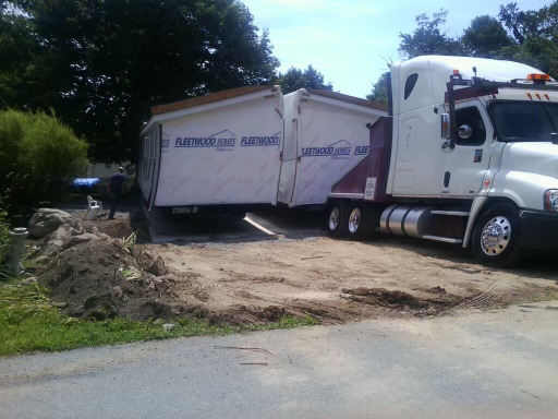 A large truck transporting a prefabricated trailer home on a dirt road, surrounded by trees.