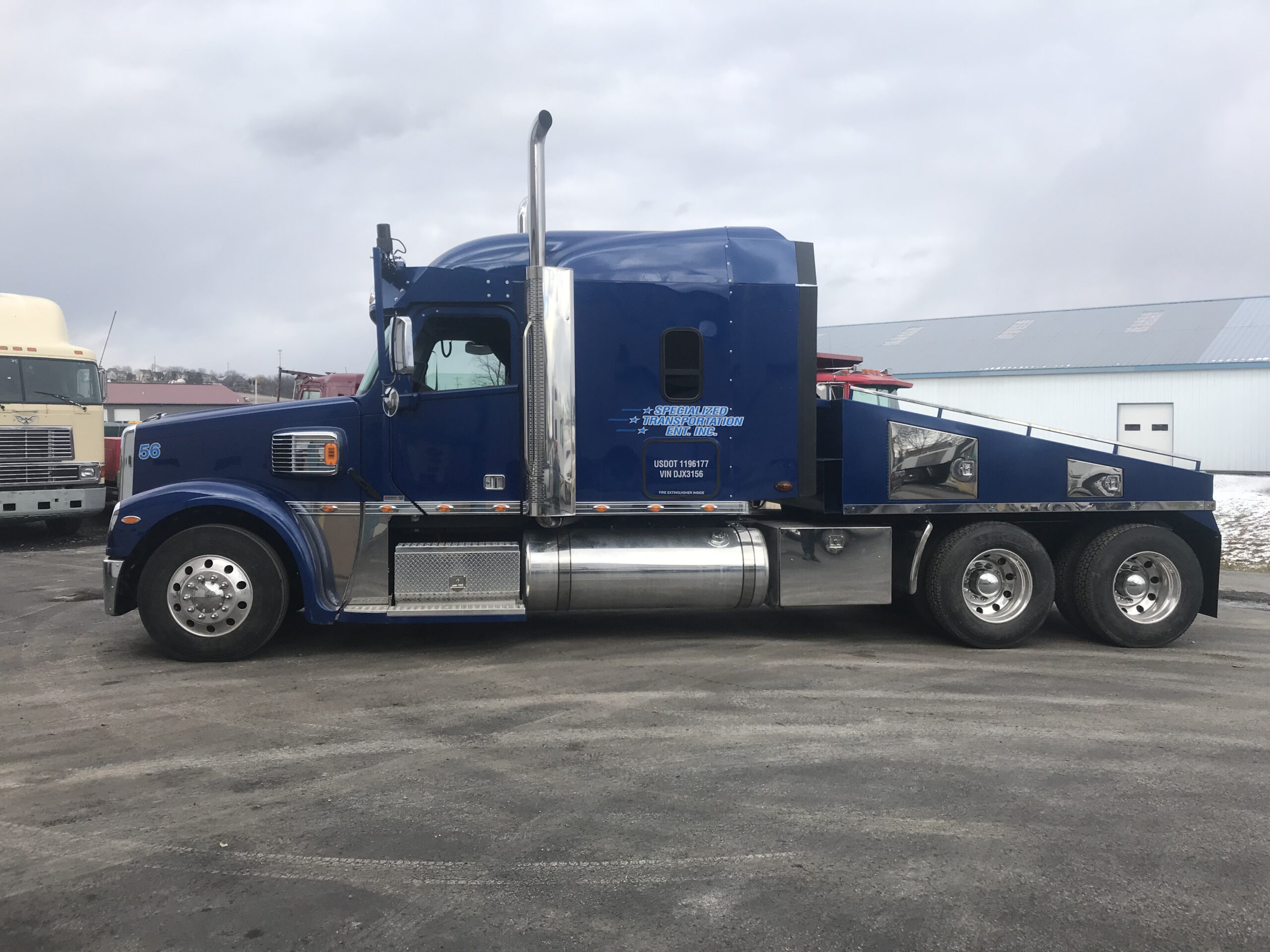 A blue semi-truck parked on a paved lot with overcast skies in the background.