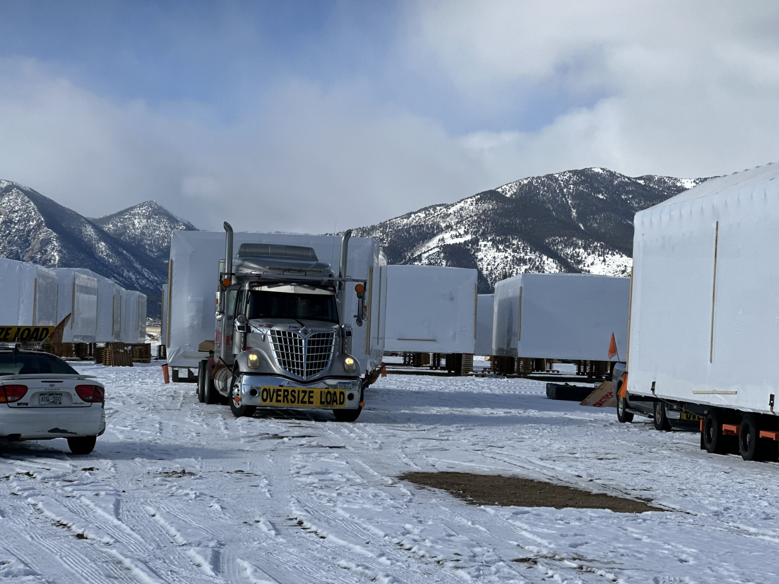Truck with "oversize load" sign transports large white structure through snowy mountain landscape.