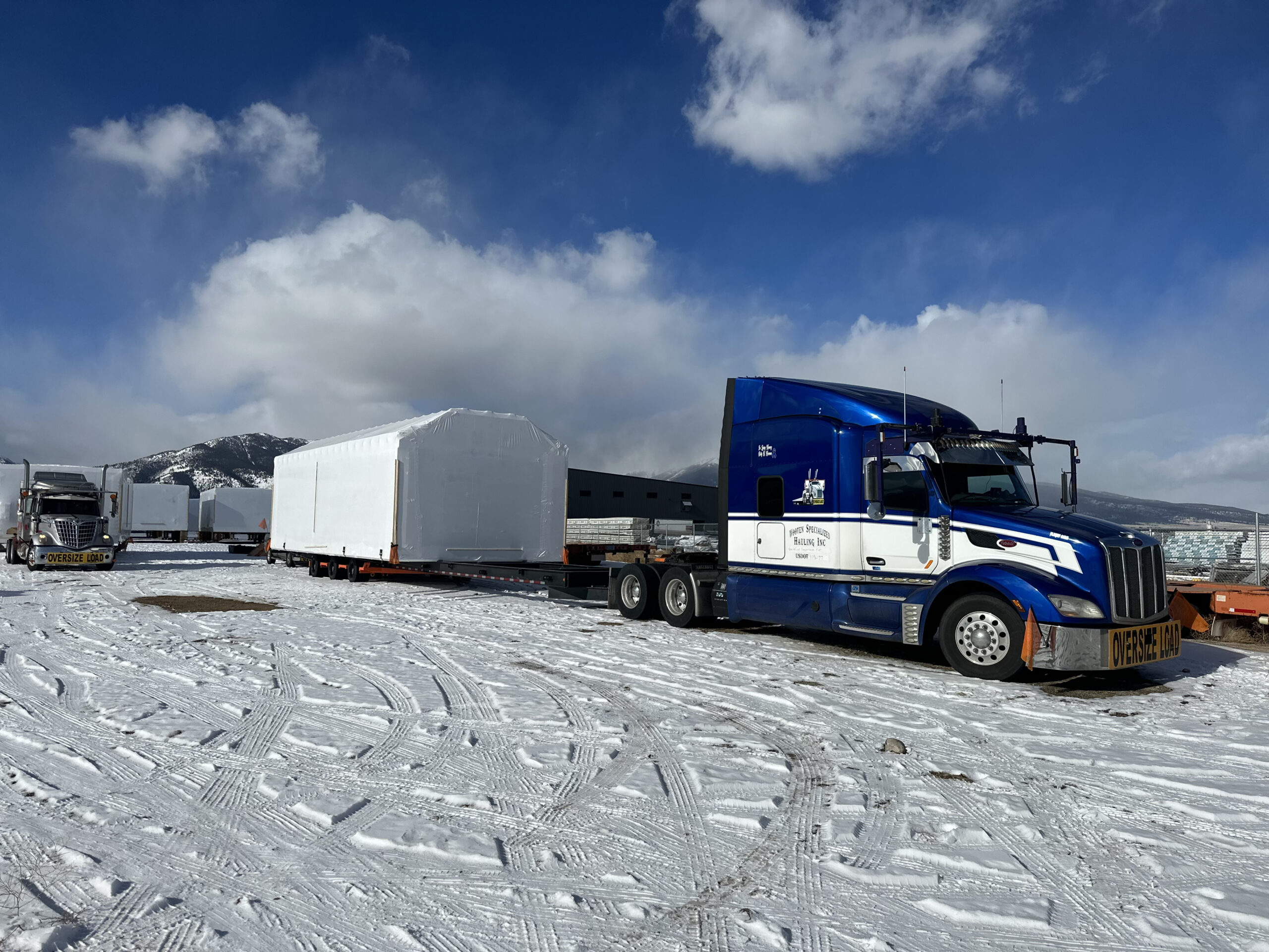 A blue semi-truck transports a large, shrouded load on a snowy landscape with mountains in the background.
