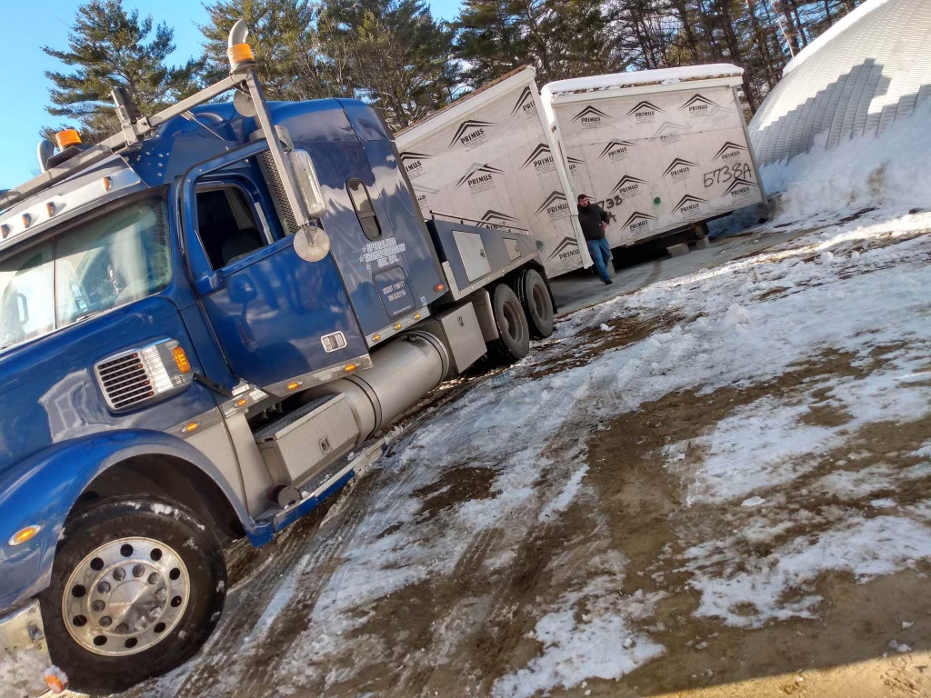 A blue semi-truck hauls a large load of building materials on a snowy, muddy site. Trees and a white structure are in the background. A person stands nearby.