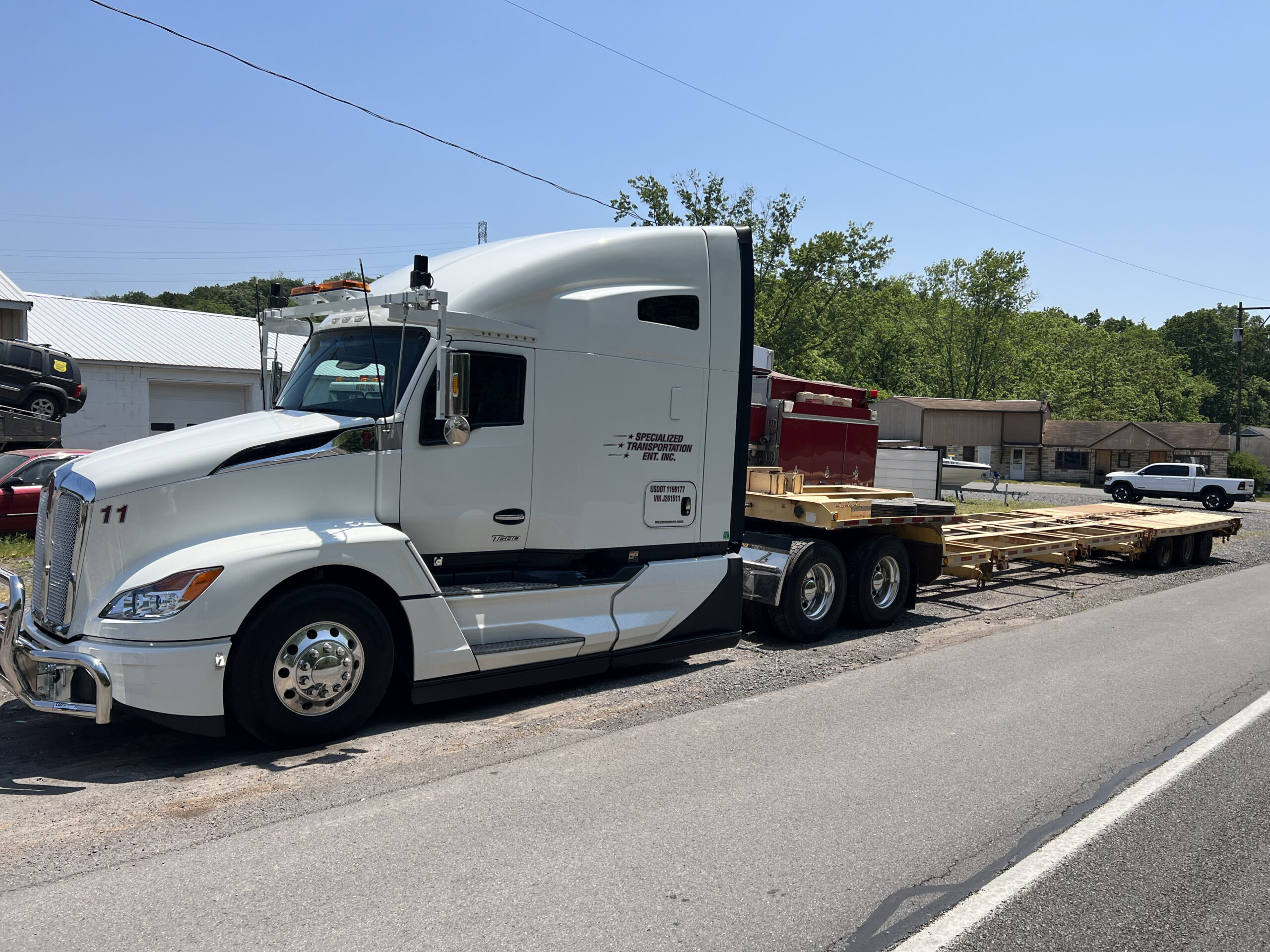 A white semi-truck is parked on the roadside, hauling a flatbed trailer loaded with wooden planks.
