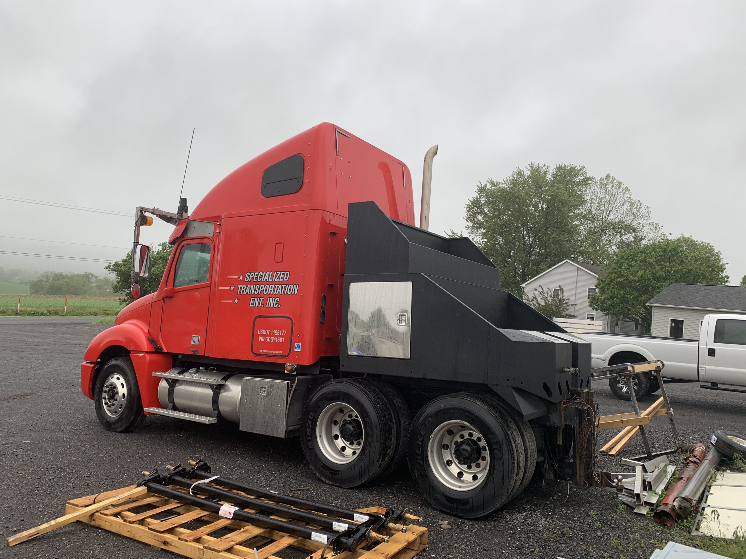Red semi-truck with black rear attachment, parked in a lot with overcast skies. Nearby are a house, trees, and a white pickup truck. Debris is visible on the ground.