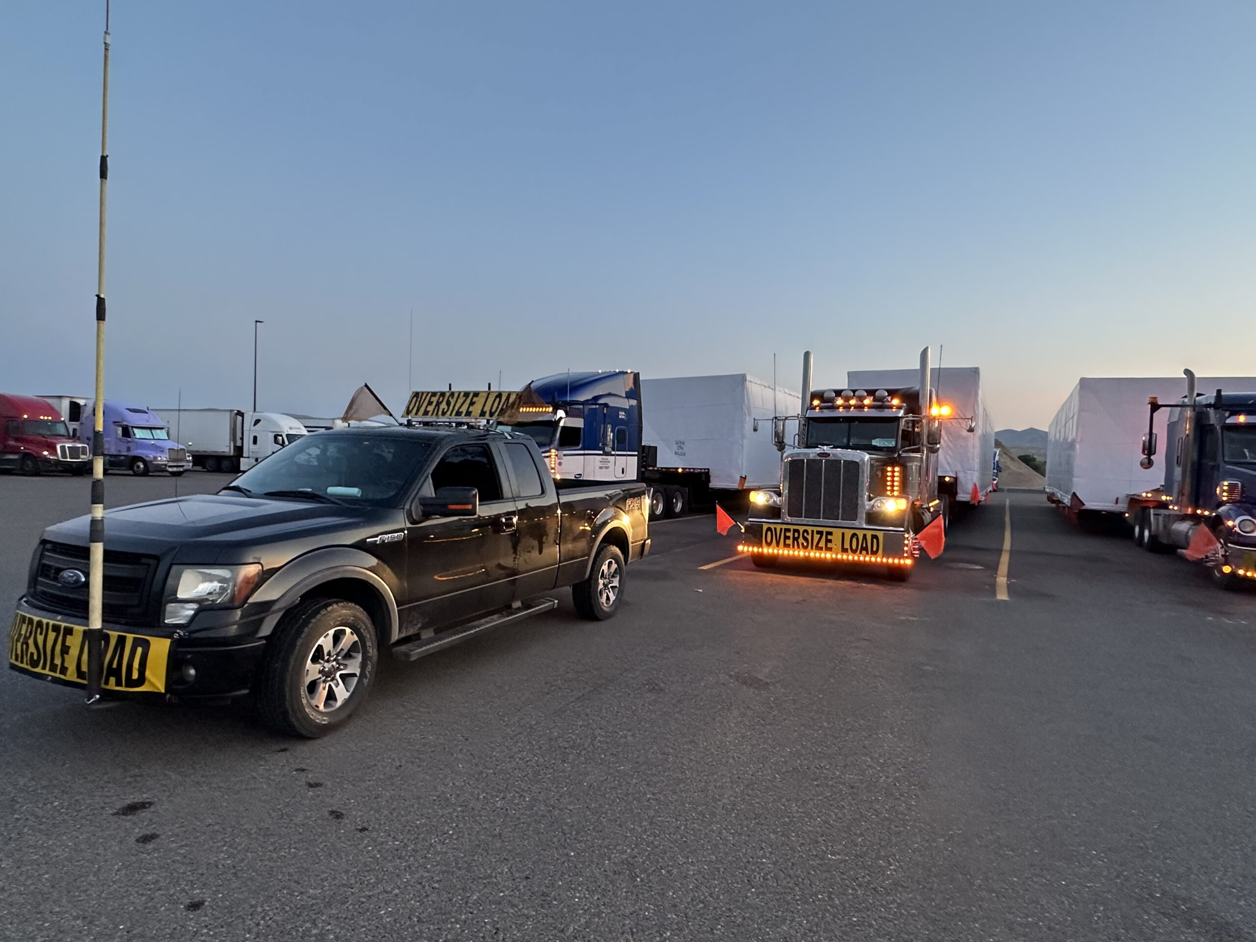 A pickup truck and a semi-truck with "Oversize Load" signs are parked in a lot with other trucks.