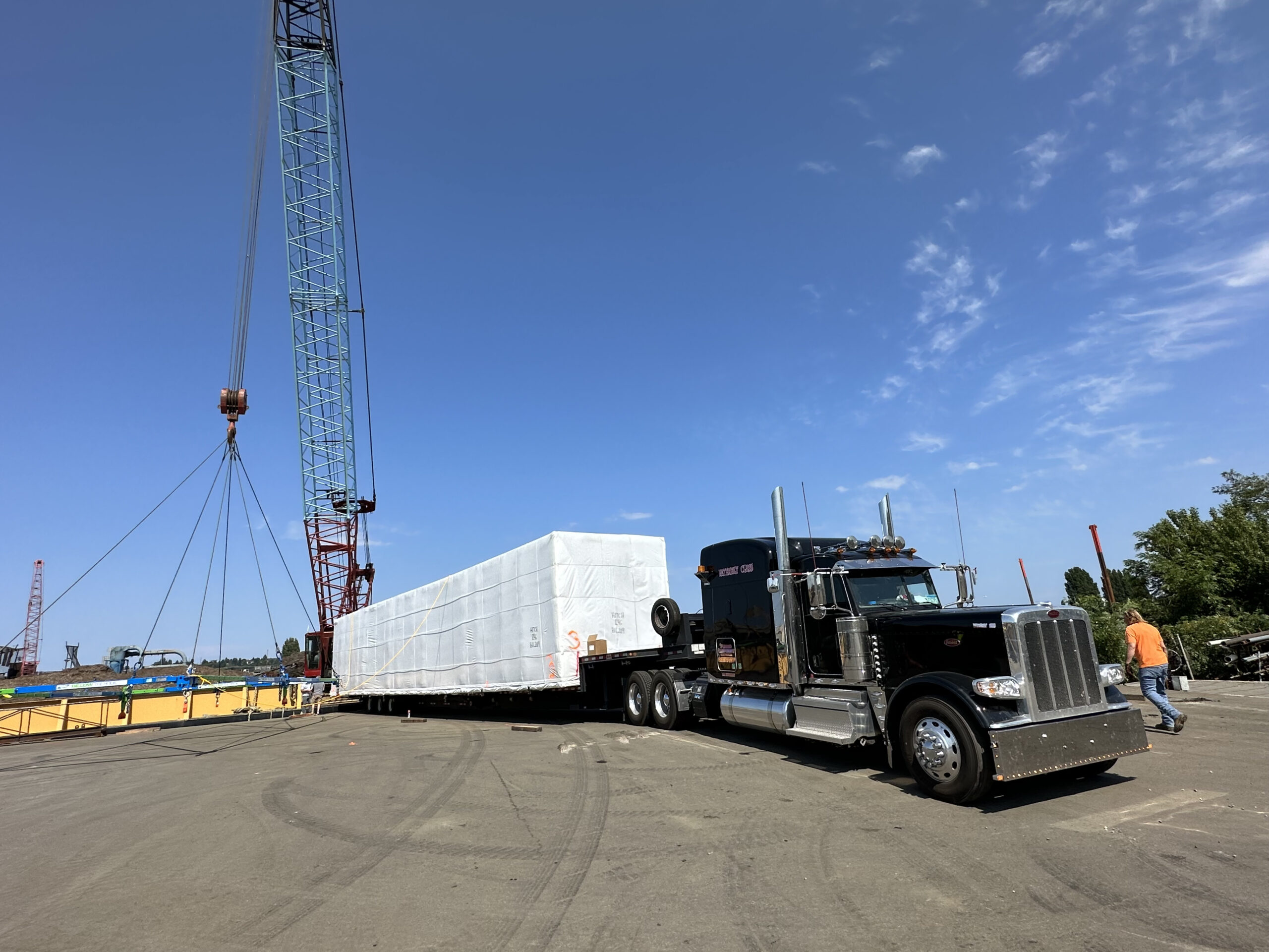 A large truck hauls a long, wrapped load at a construction site with a tall crane against a clear blue sky.