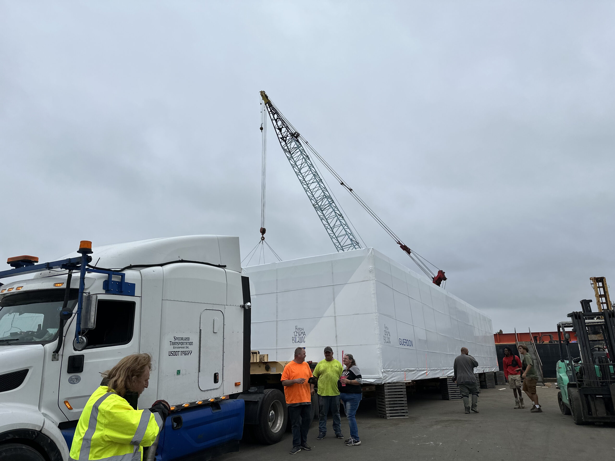 A truck with a large white container, people in safety vests standing nearby, and a crane in the background under an overcast sky.