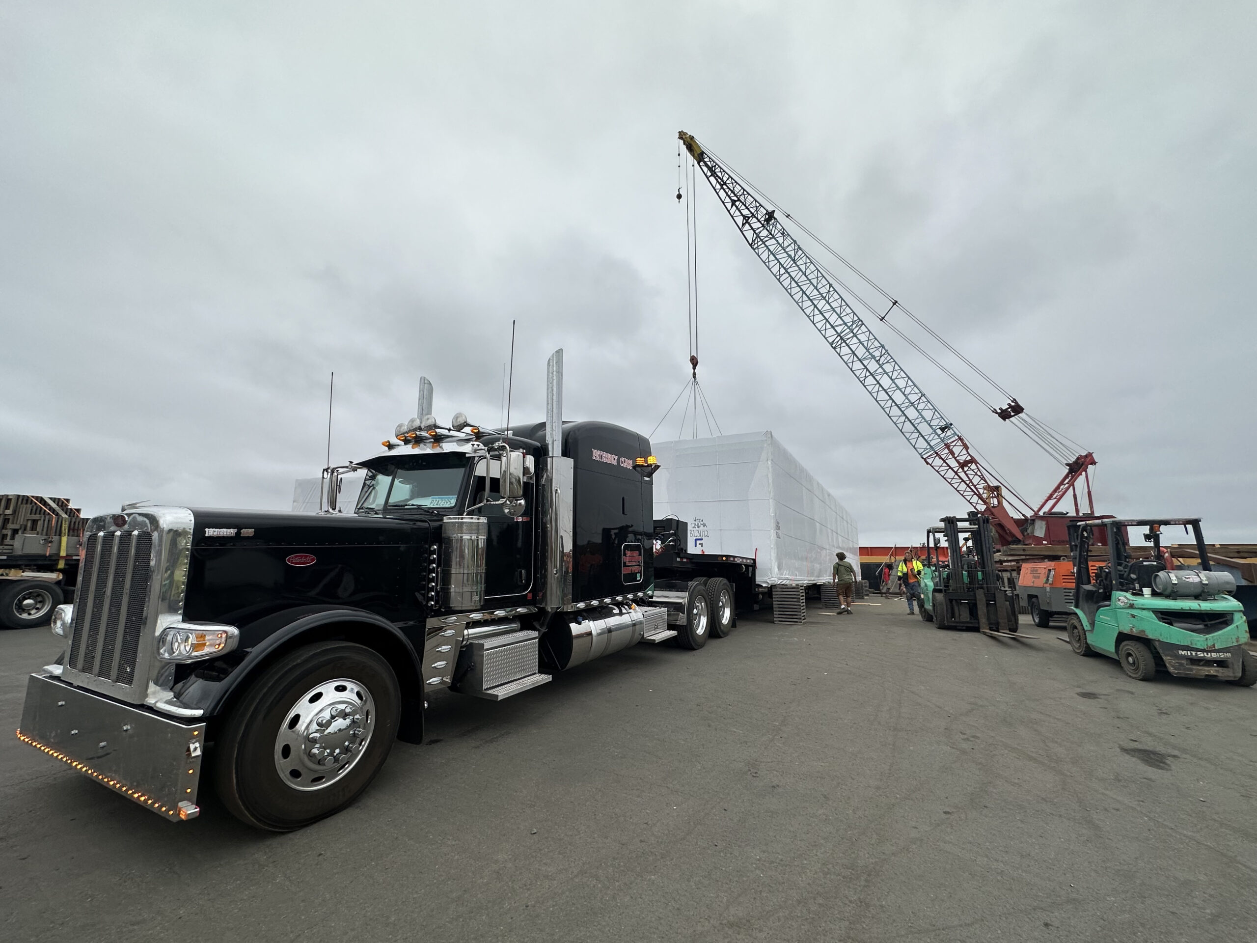 A large black truck is parked on a paved surface, with a crane lifting a covered object nearby. Several people and forklifts are visible in the background under a cloudy sky.