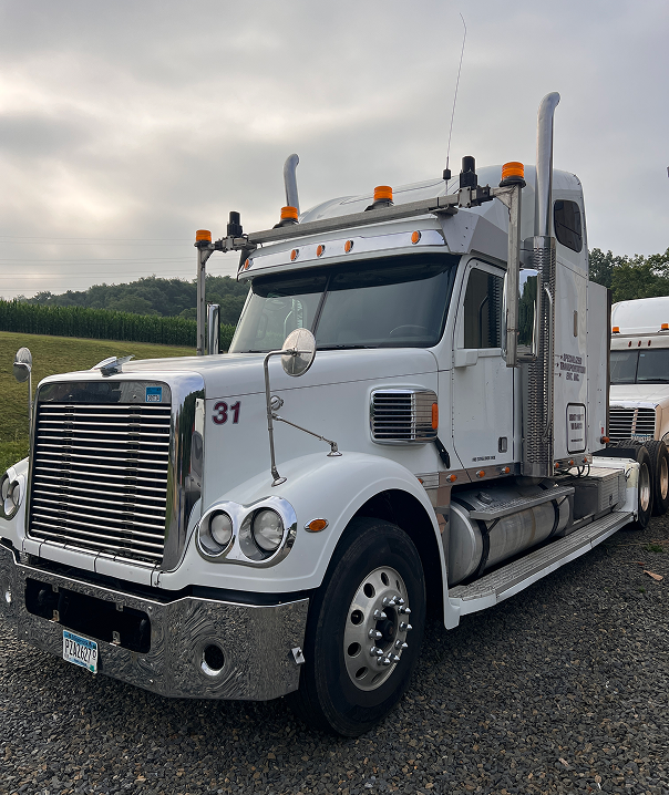 A white semi-truck with chrome trim and orange lights is parked on gravel beside a field, with another truck partially visible in the background.
